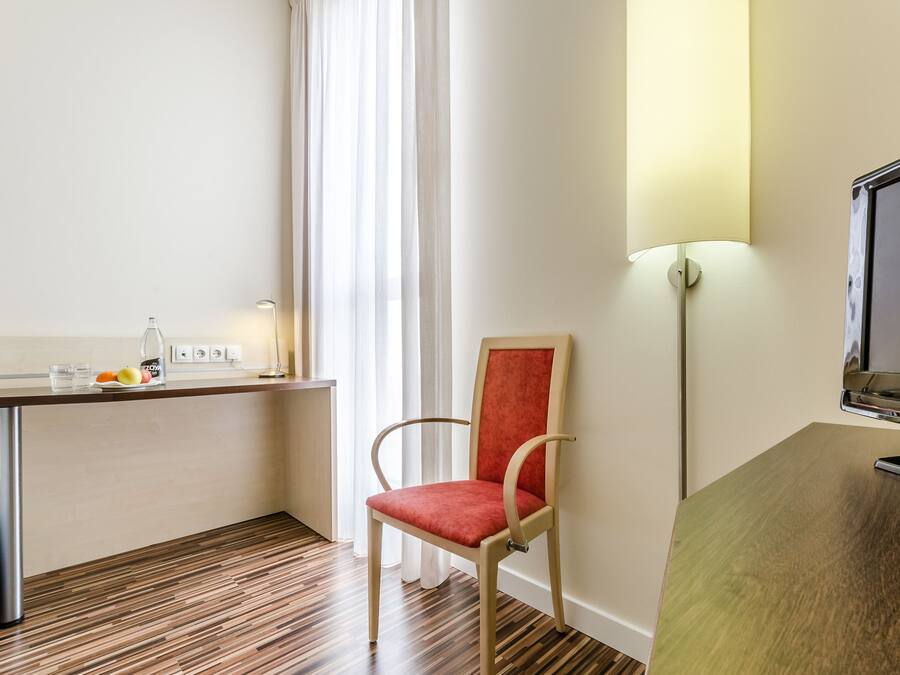 A hotel room with a wooden floor, a desk with a lamp, a chair, and a TV on a stand.