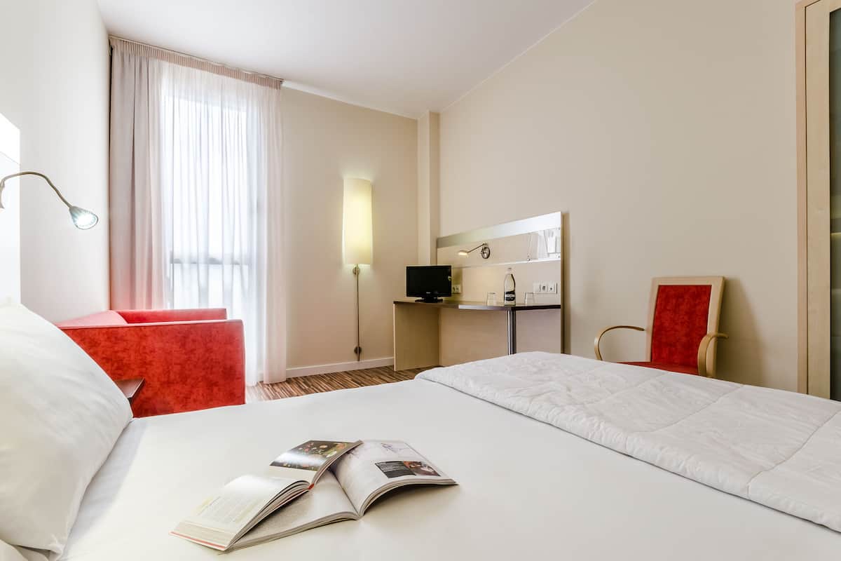 A hotel room with a large bed, a red armchair, a desk with a computer, and a TV.