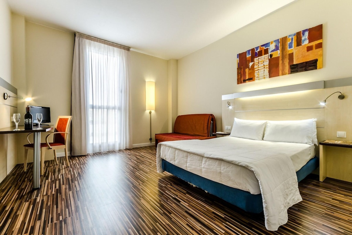 A hotel room with a bed, a desk with a computer, a chair, a window with curtains, and a painting on the wall.