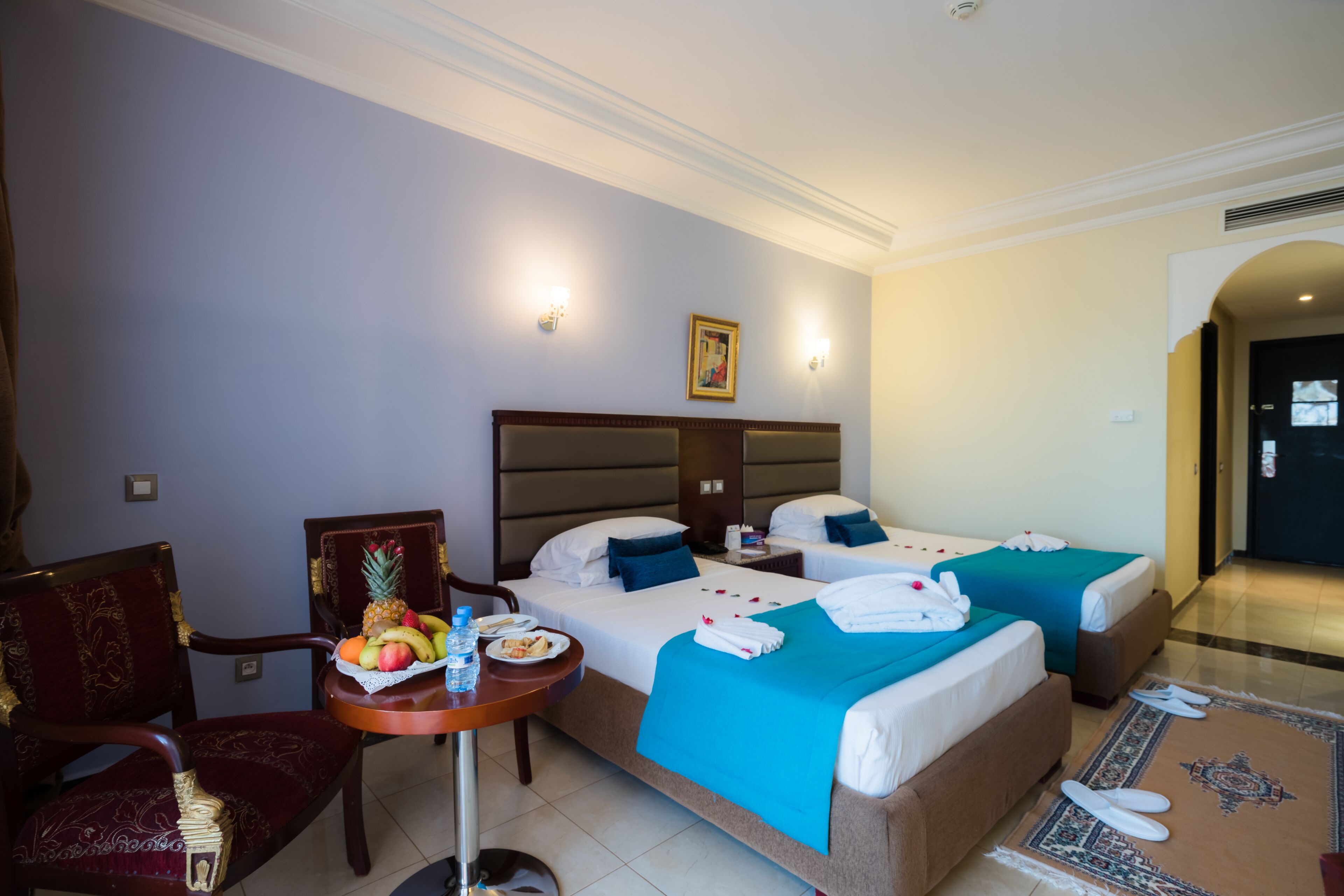 Habitación de hotel con dos camas, una mesa de comedor con fruta y un plato de comida, una silla y una mesita con una botella de agua.