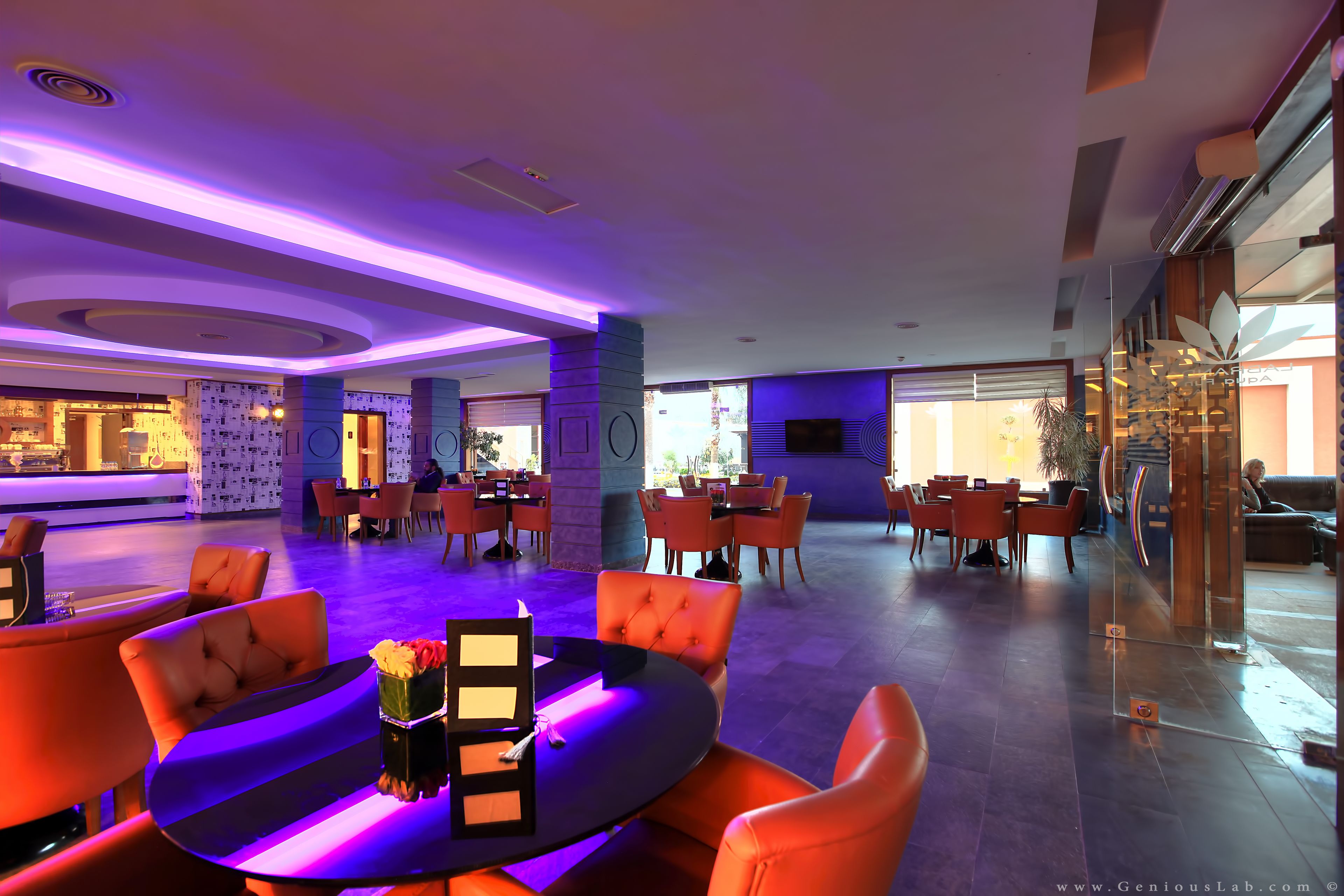Un hall d’hôtel moderne avec une table ronde centrale, des sièges orange et un éclairage d’ambiance violet.