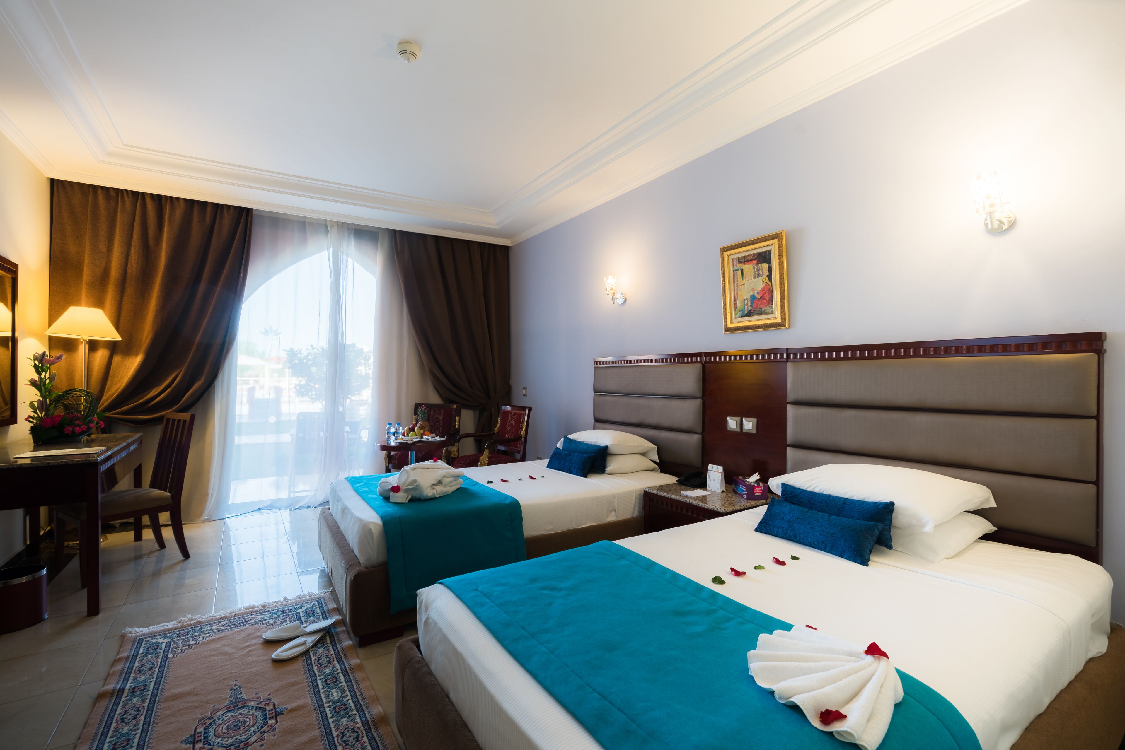 A hotel room with two beds, a desk, a chair, a lamp, and a painting on the wall.