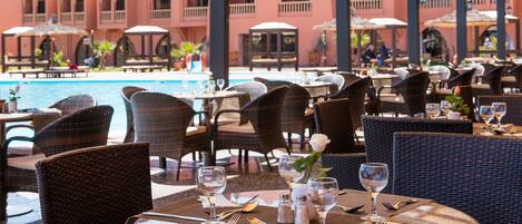 Un restaurante junto a la piscina con mesas dispuestas para comer, una piscina y un edificio con balcones al fondo.