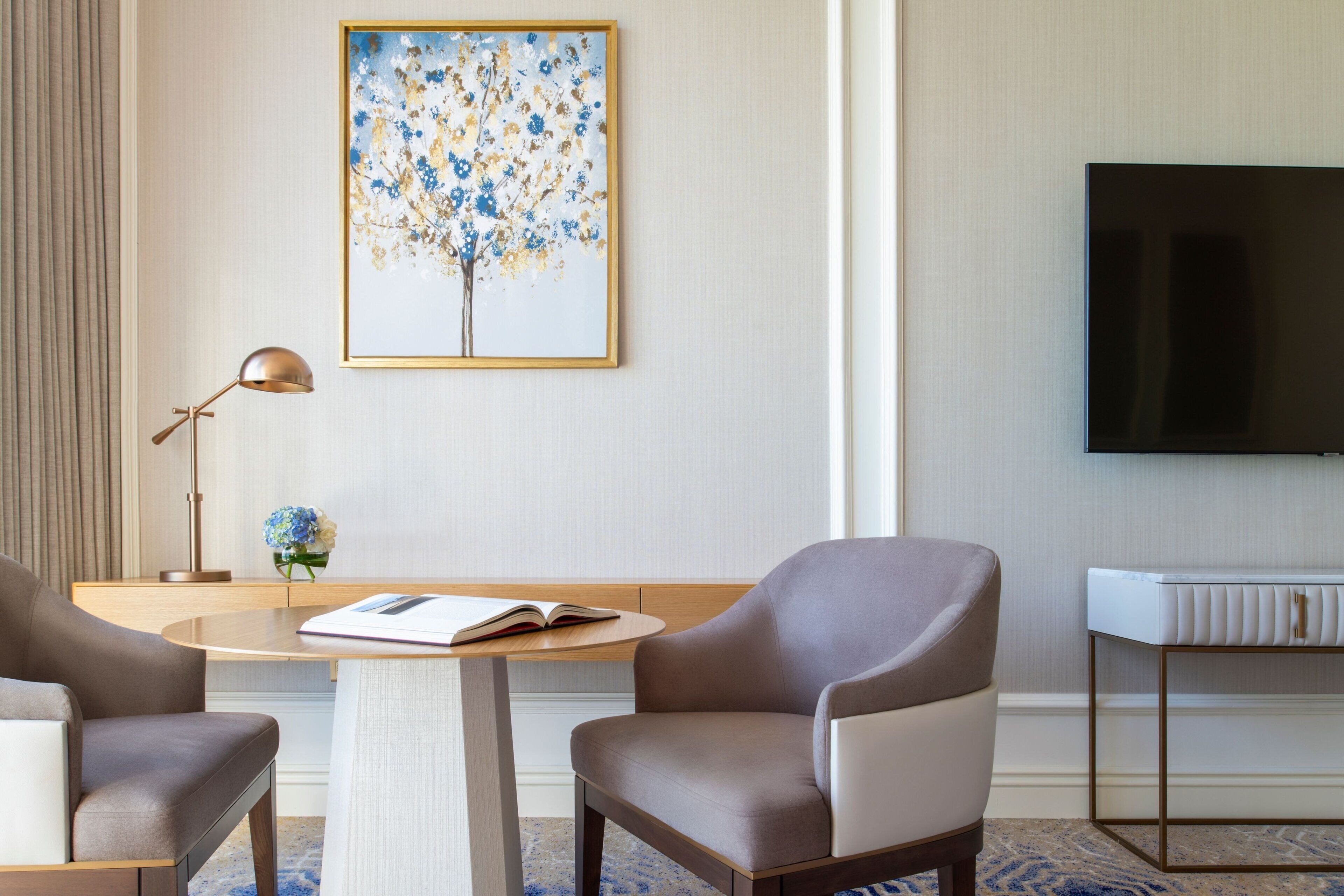 Une chambre d’hôtel moderne avec une table ronde, deux fauteuils, un téléviseur, une œuvre d’art encadrée et une table de chevet.