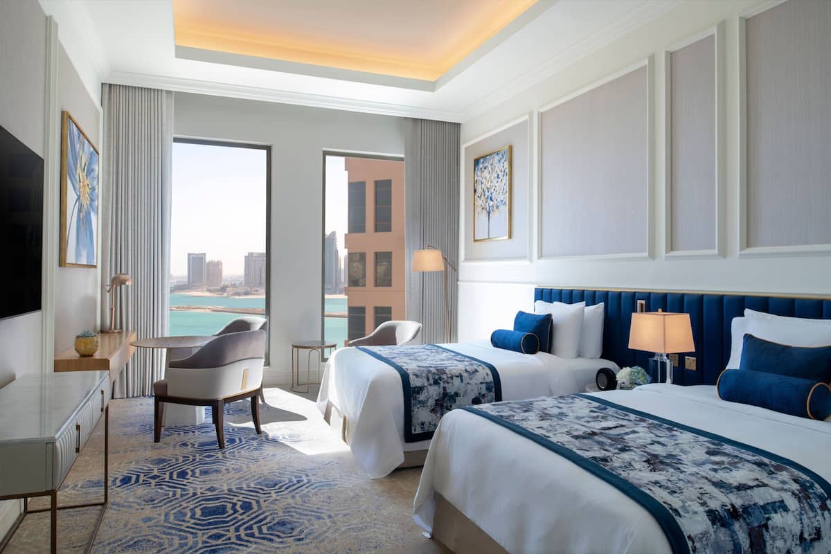 Een hotelkamer met twee bedden, een bureau, een stoel en een groot raam met uitzicht over de stad.