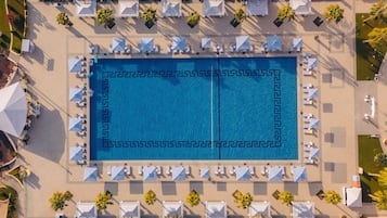 An aerial view of a resort with a central swimming pool, surrounded by lounge areas with umbrellas and sun loungers, and palm trees.