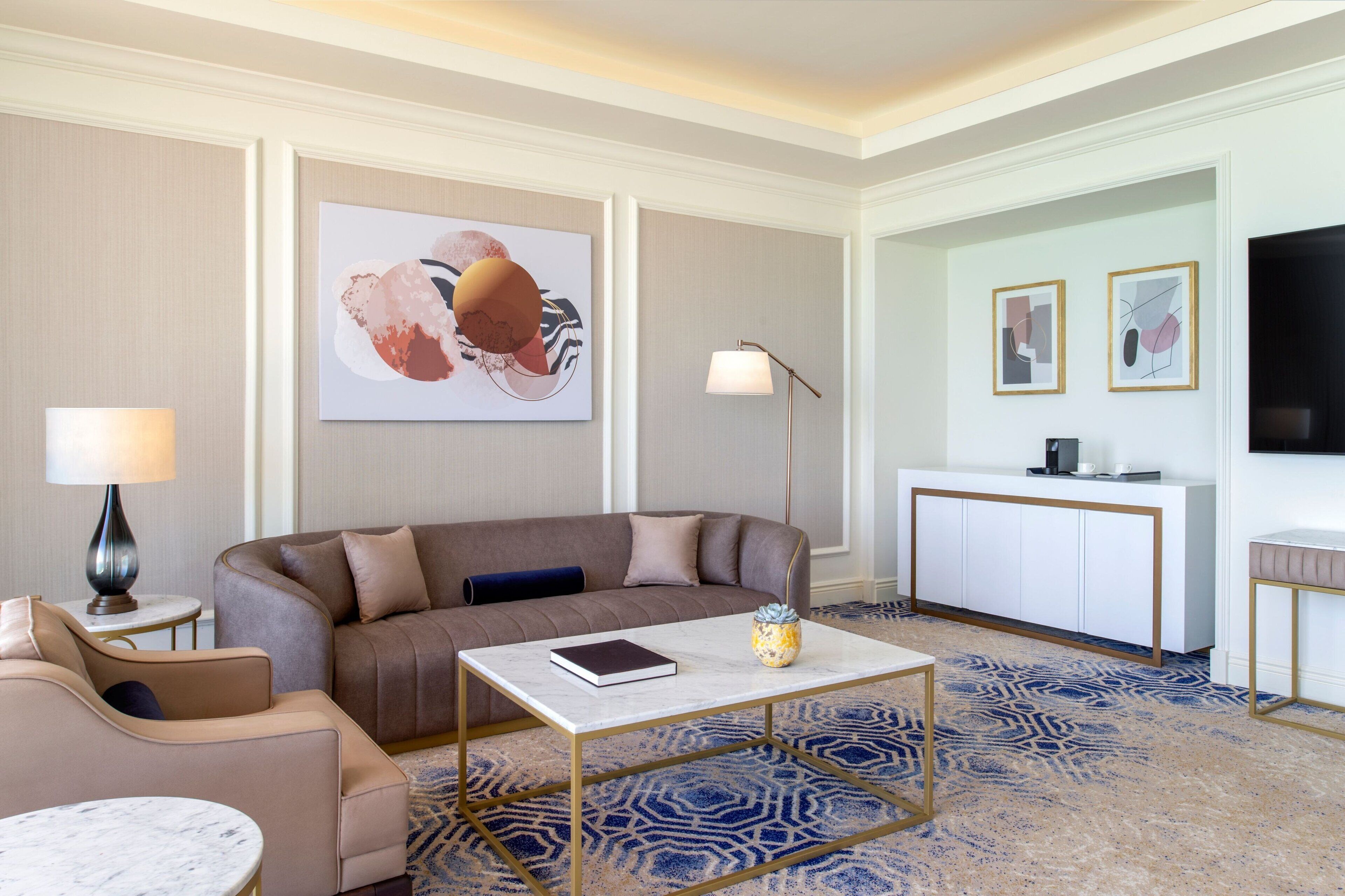A modern hotel room with a sofa, a small table, a lamp, a coffee table, a desk, and artwork on the walls.