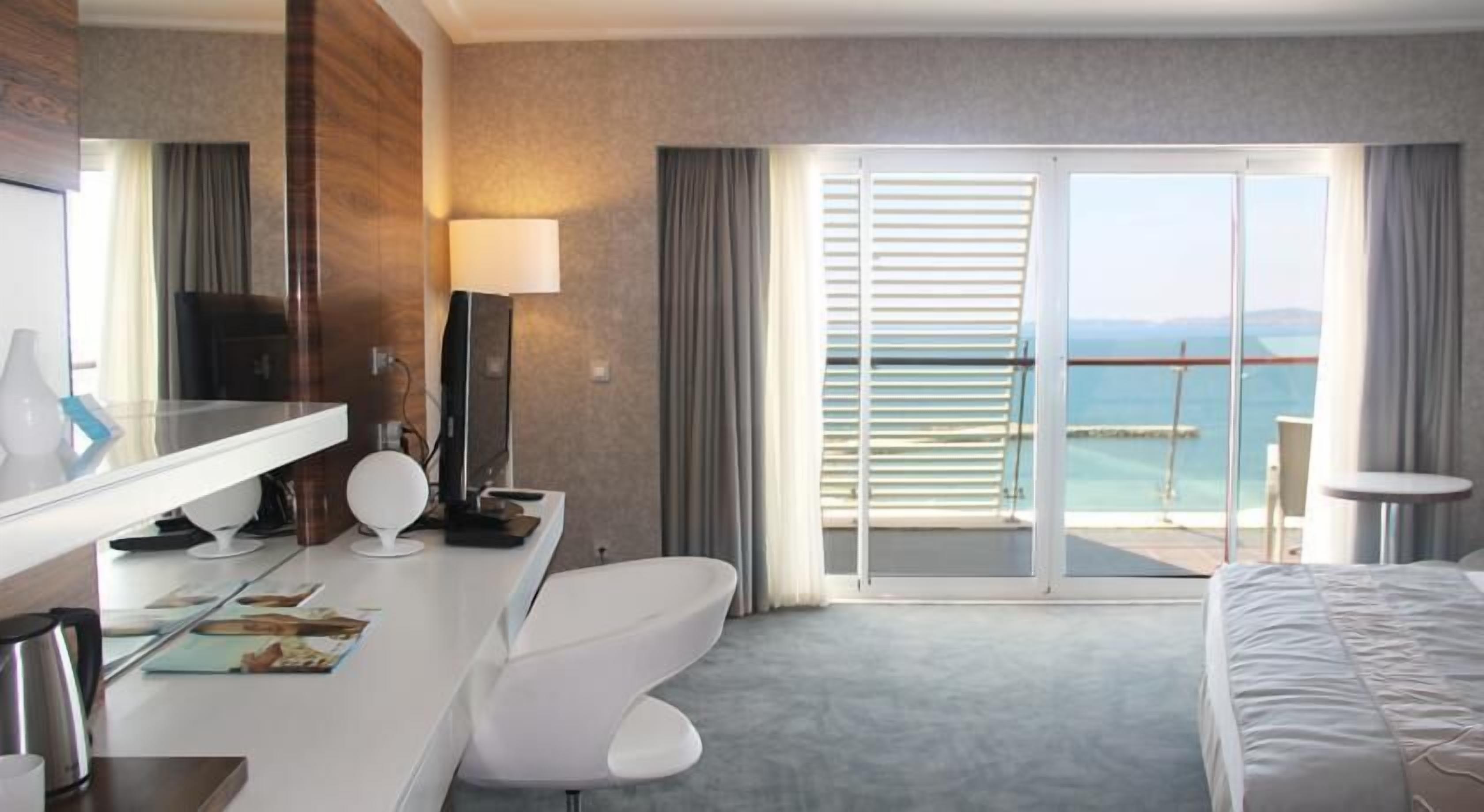 Ein Hotelzimmer mit Bett, Schreibtisch mit Computer, Stuhl, großem Spiegel und Meerblick durch eine Schiebetür.