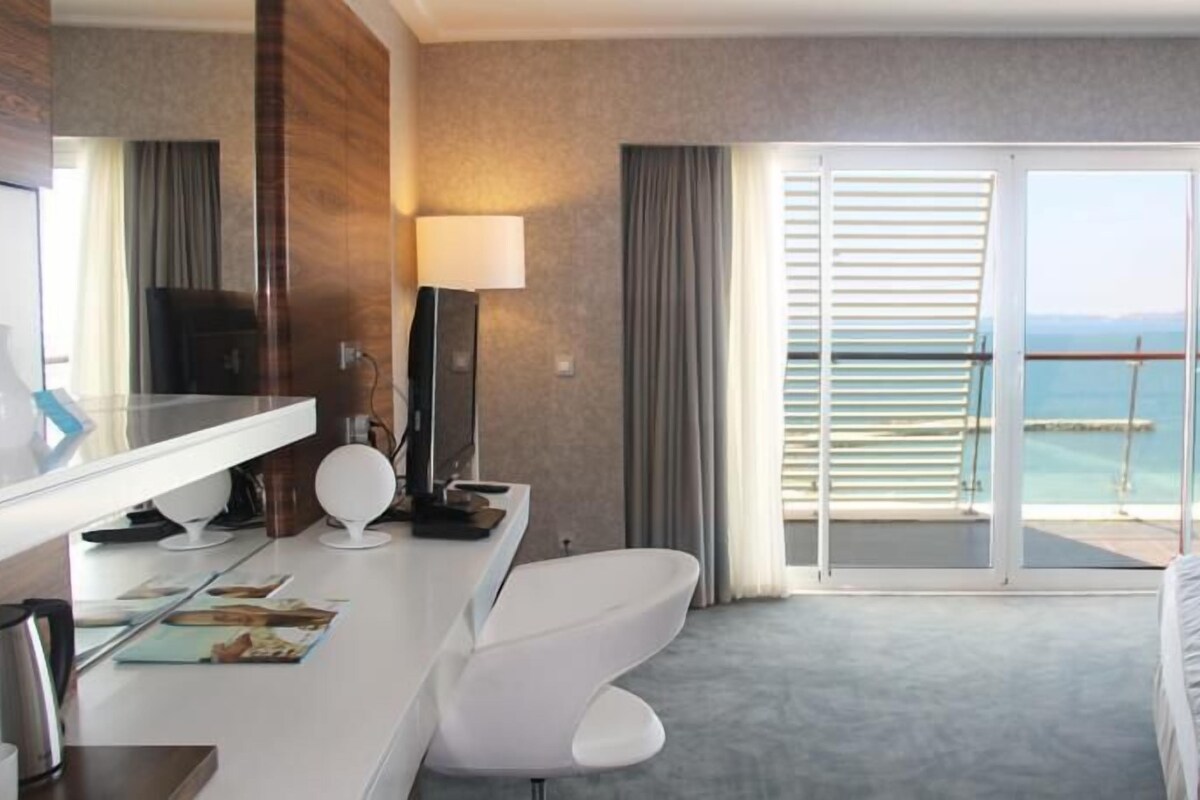 A hotel room with a bed, a desk with a computer, a chair, a large mirror, and a view of the sea through a sliding glass door.
