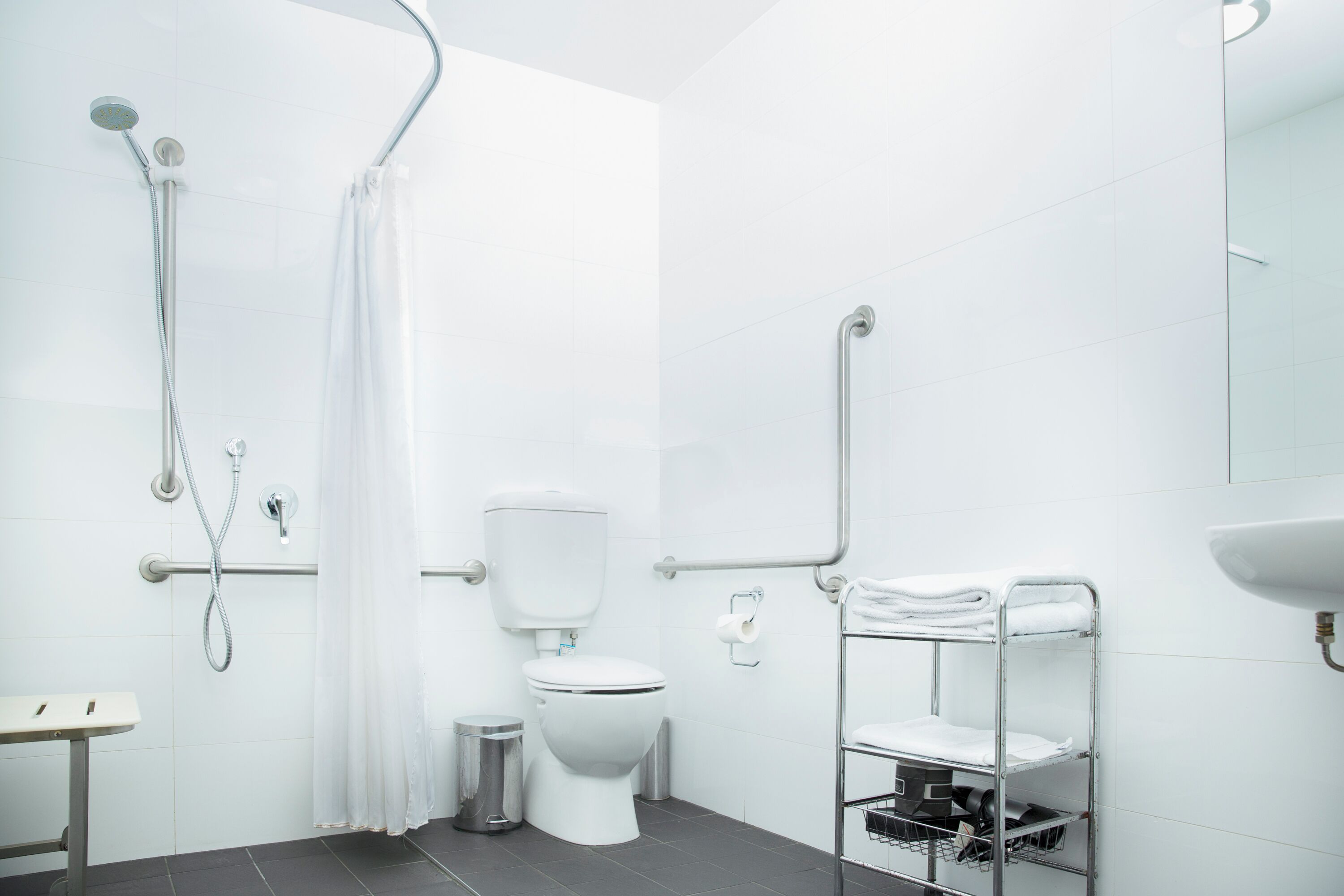 A modern bathroom with a white toilet, a shower with a curved hose, a grab bar, a sink, and a towel rack.