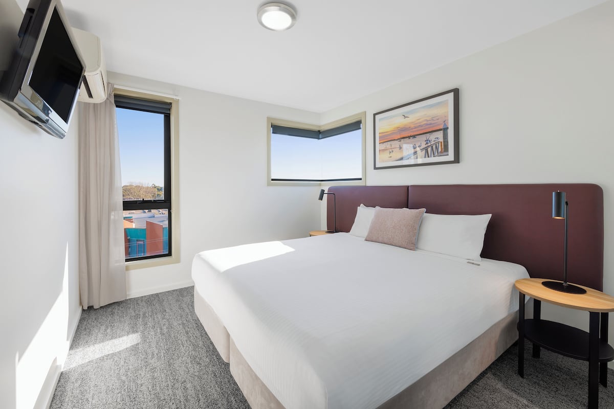 Ein Hotelzimmer mit einem großen Bett, einem an der Wand montierten Fernseher, einem Fenster mit Blick auf Gebäude und einem gerahmten Bild an der Wand.