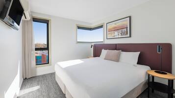 A hotel room with a large bed, a TV mounted on the wall, a window with a view of buildings, and a framed picture on the wall.
