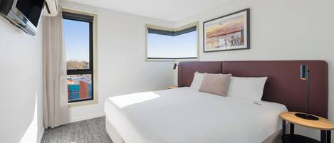 A hotel room with a large bed, a TV mounted on the wall, a window with a view of buildings, and a framed picture on the wall.