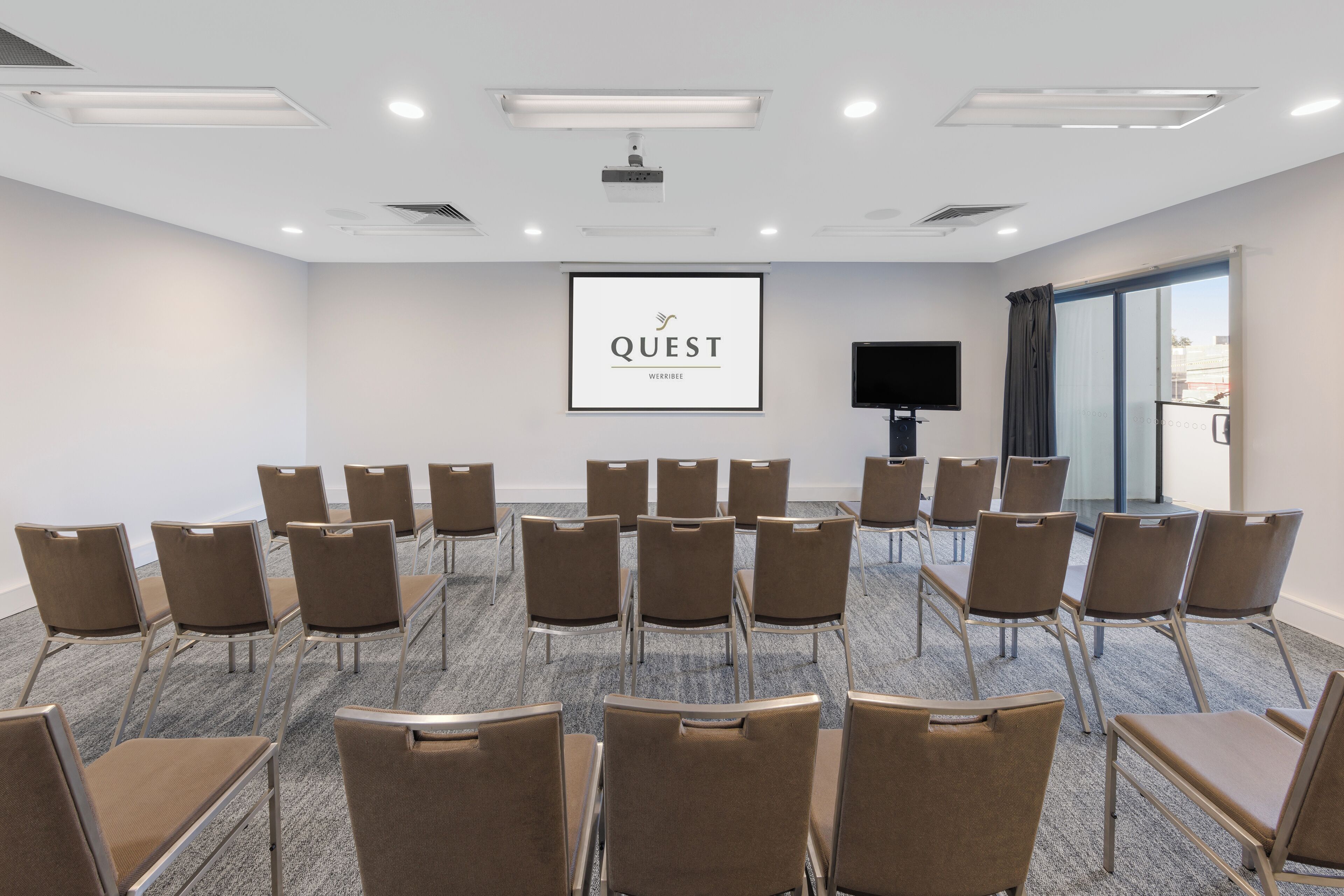 Un salón de conferencias con filas de sillas, una pantalla de proyector y un televisor.