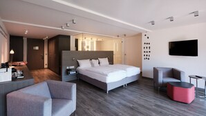 A modern hotel room with a large bed, a desk, a chair, a TV, and a small table.