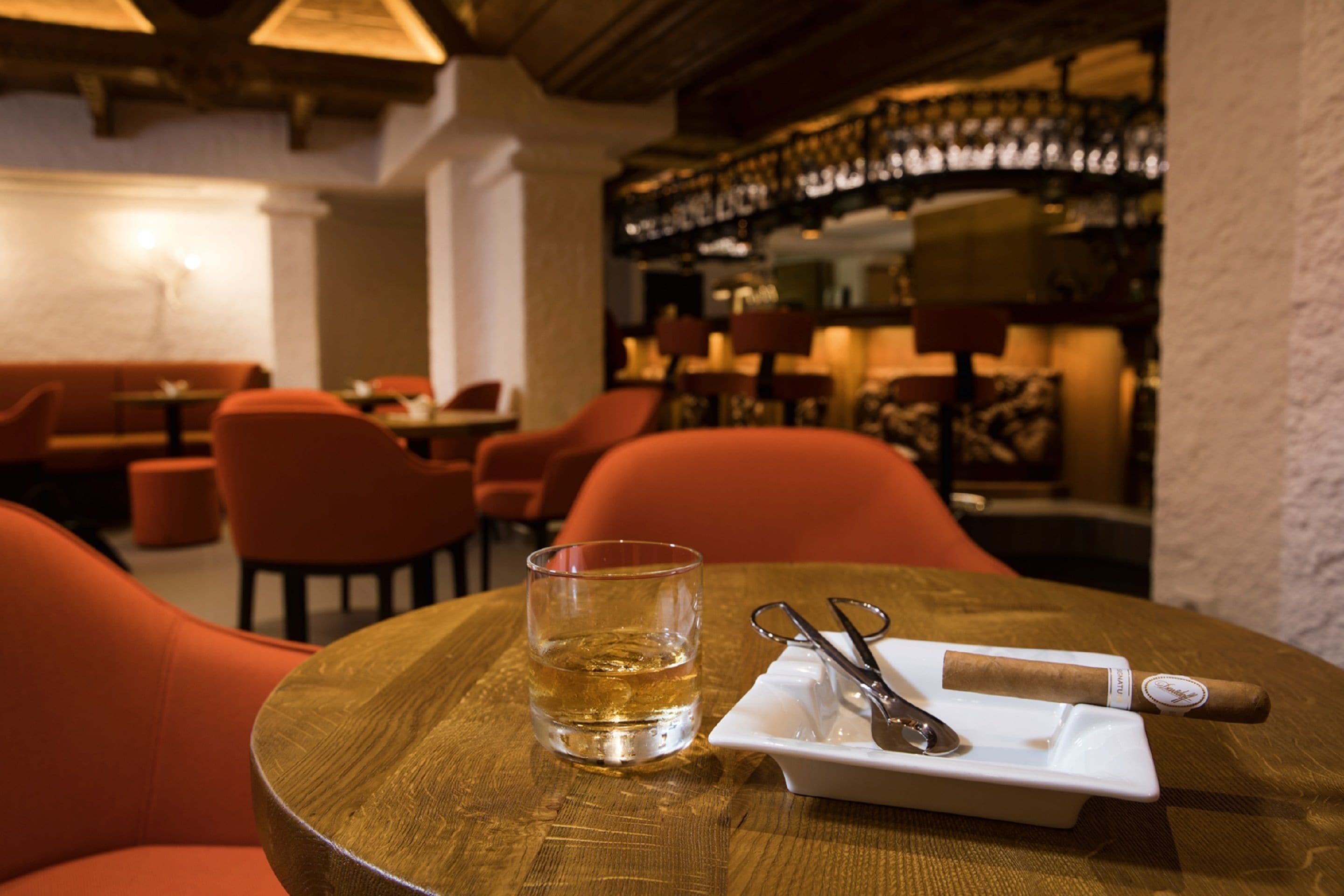A round wooden table with a glass of whiskey, a pair of scissors, and a cigar. The setting appears to be a restaurant with orange chairs and a bar in the background.