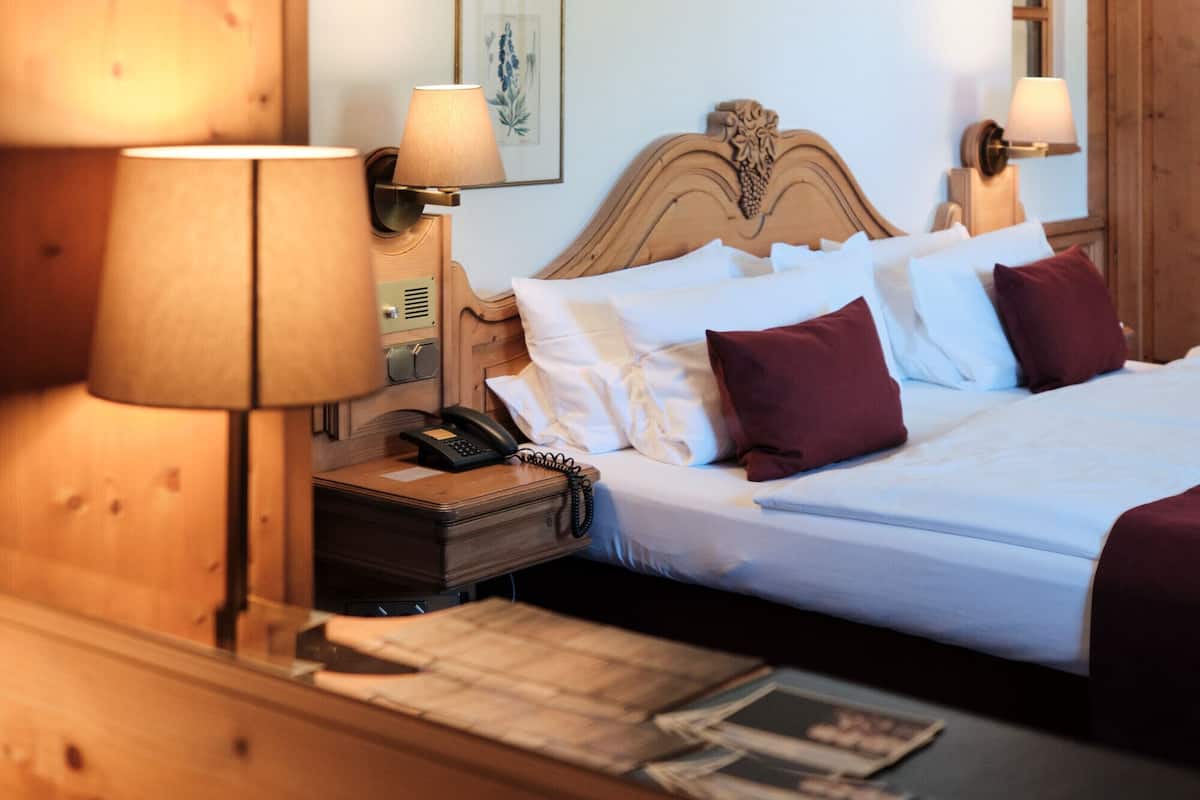 Ein Hotelzimmer mit hölzernem Kopfteil, zwei Nachttischlampen, einem Telefon, einem Nachttisch mit Lampe und einem Bett mit weißer und bordeauxroter Bettwäsche.