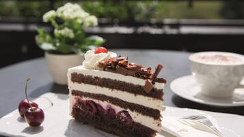 Une part de gâteau au chocolat, composé de plusieurs couches, garnie de crème fouettée et de cerises, présentée sur une assiette blanche, accompagnée d’une tasse de café posée sur une soucoupe.