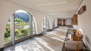 Un pasillo amplio con ventanas arqueadas grandes, una silla de madera y vistas a un jardín y montañas.