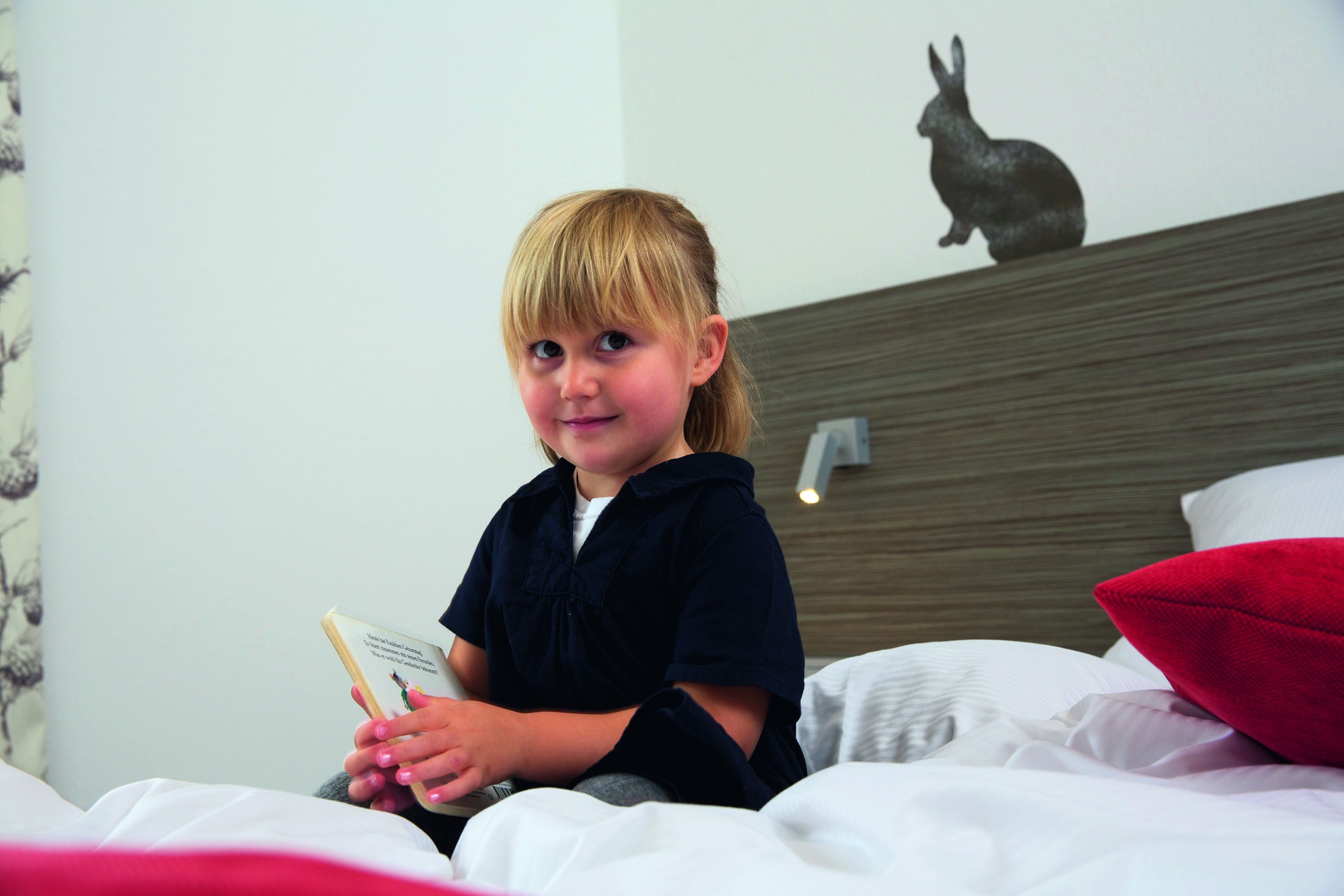 A child sitting on a bed with a book, a red pillow, and a wall-mounted rabbit sculpture.