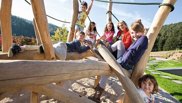 Eine Gruppe Kinder spielt auf einer hölzernen Spielanlage mit einer Seilbrücke vor einer bewaldeten Landschaft.
