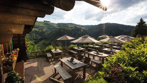 Un patio al aire libre con mesas y sillas de madera, sombrillas y vistas a un valle boscoso.