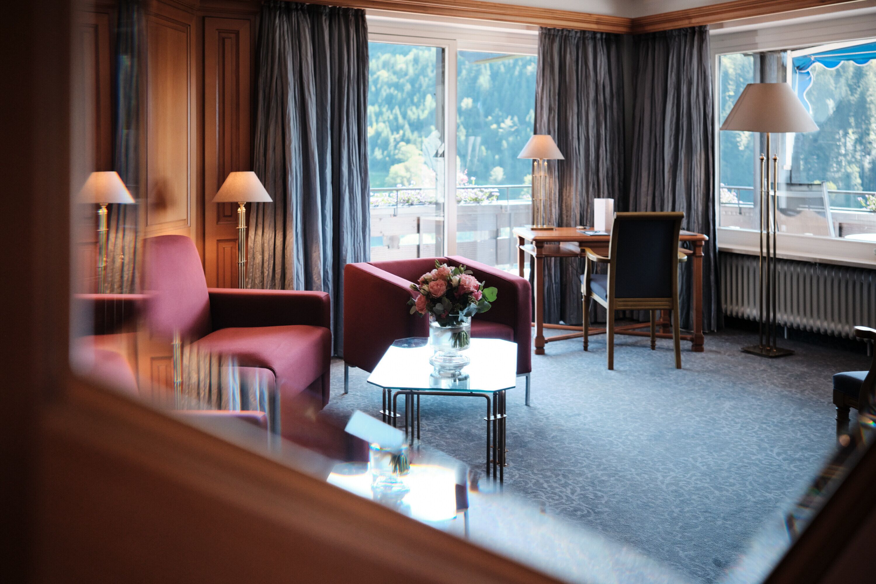 A hotel room with a large window, a red armchair, a glass coffee table with a vase of flowers, and two floor lamps.