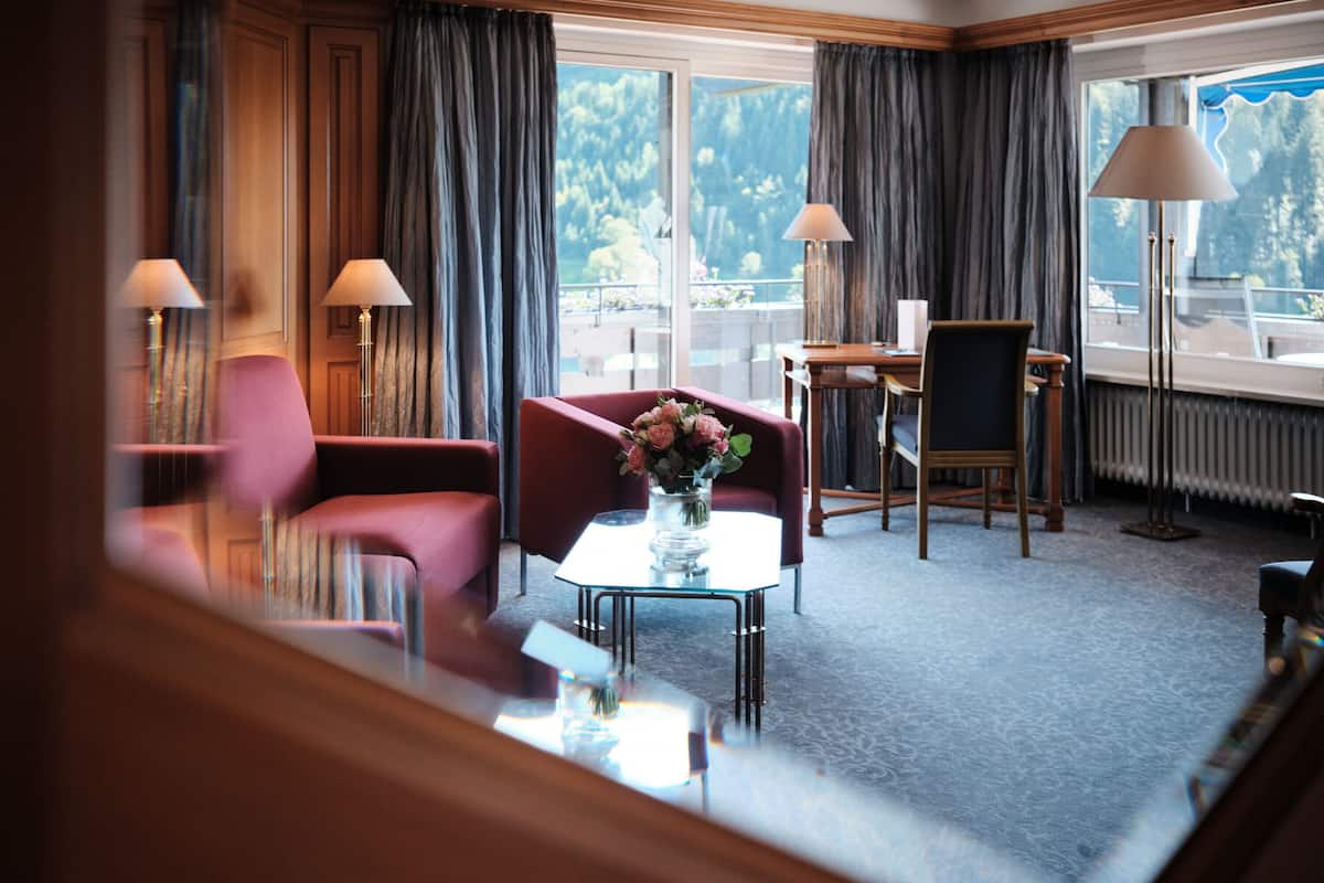 Ein Hotelzimmer mit großem Fenster, einem roten Sessel, einem Glastisch mit Blumenvase und zwei Stehlampen.