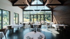 Un comedor con mesas redondas, sillas y grandes ventanales que ofrecen vistas a un paisaje boscoso.