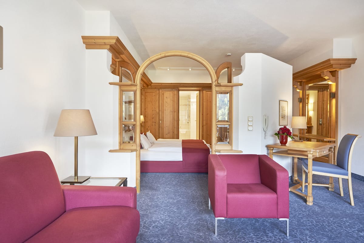 Ein Hotelzimmer mit einem Bett, zwei roten Sessel, einem Holztisch mit Stuhl, einer Lampe und einem Spiegel.