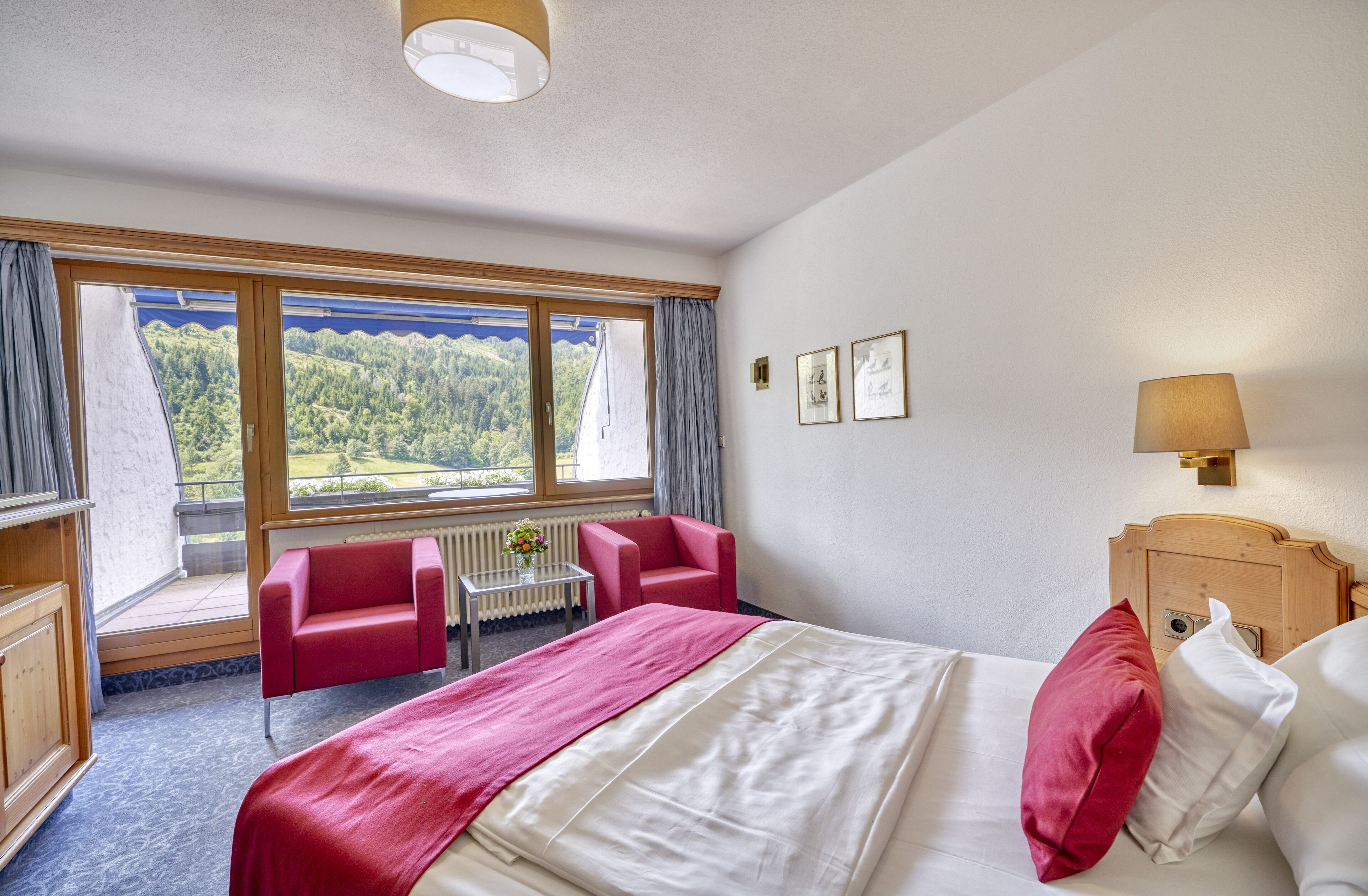 Een hotelkamer met een groot bed, een rode stoel, een kleine tafel met bloemen en uitzicht op een bos door het raam.