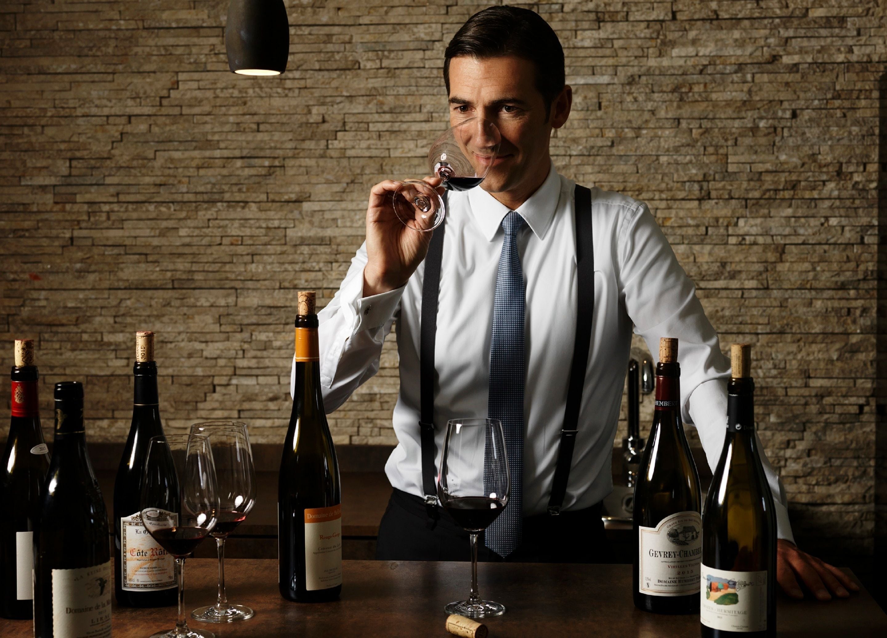 A man in a white shirt and tie holding a wine glass, standing in front of a brick wall with several wine bottles and glasses on a table.