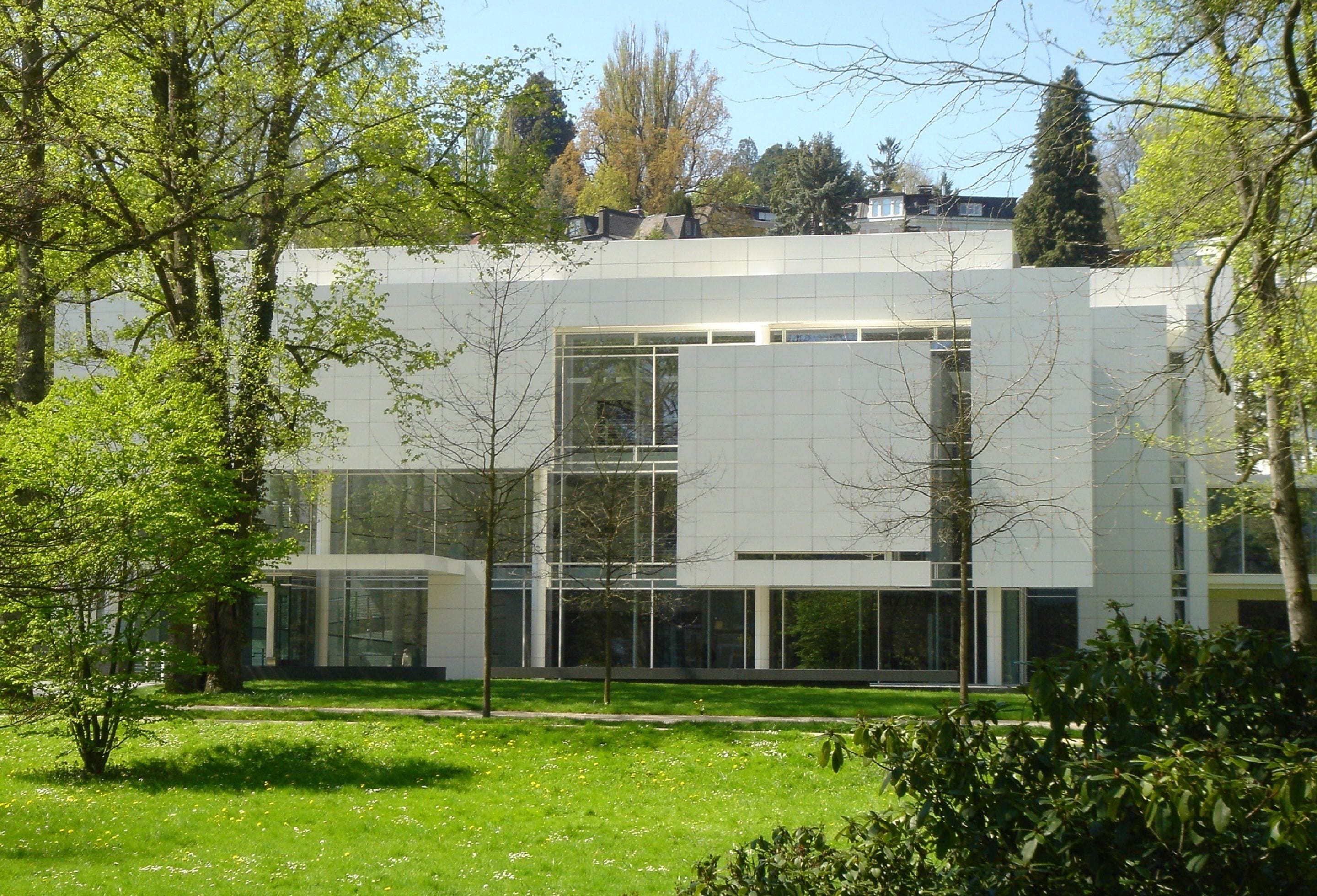Ein modernes Gebäude mit großen Glasfenstern und einem Flachdach, umgeben von einer Grünfläche mit Bäumen.