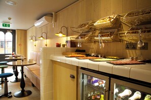 A restaurant with a counter displaying food, a seating area with tables and chairs, and a refrigerator stocked with beverages. - The Z Hotel Soho (London)