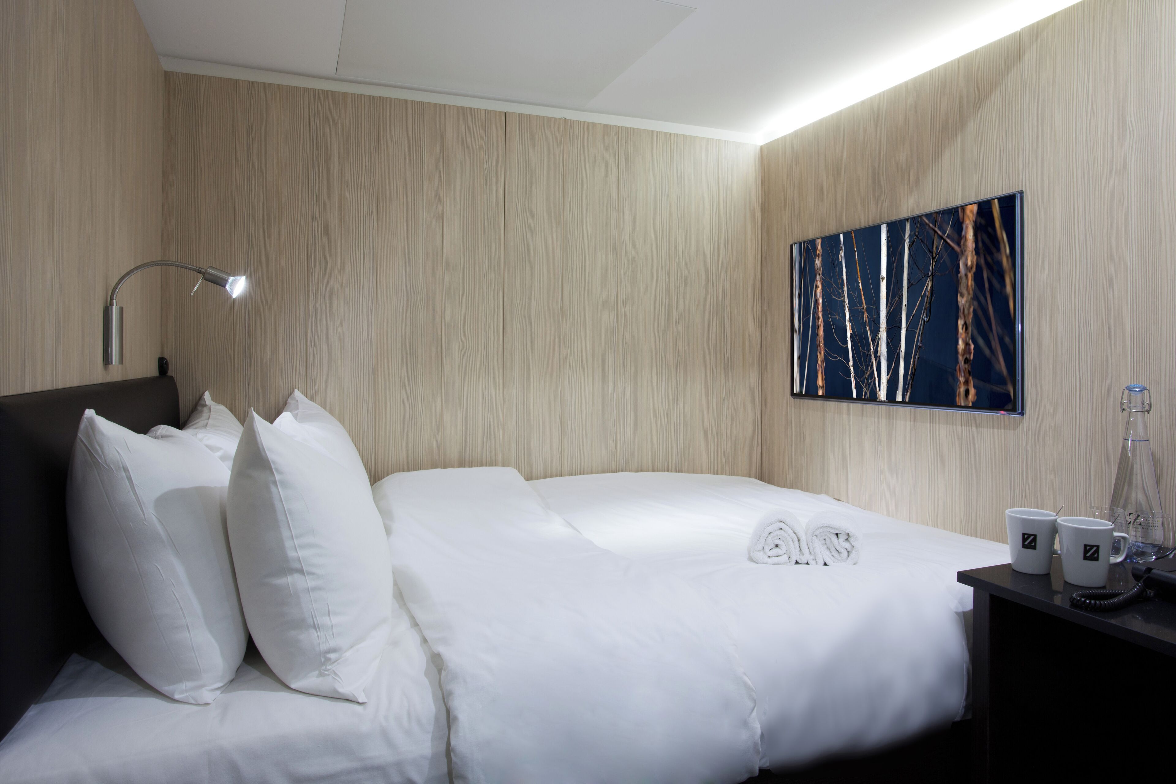 Une chambre d’hôtel avec un lit, des oreillers blancs, une table de chevet avec une lampe, un tableau au mur et une petite table de nuit avec un téléphone et deux tasses.