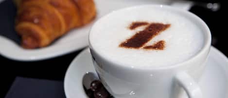 A cup of coffee with a frothy top and a chocolate powder design, accompanied by a croissant on a plate.