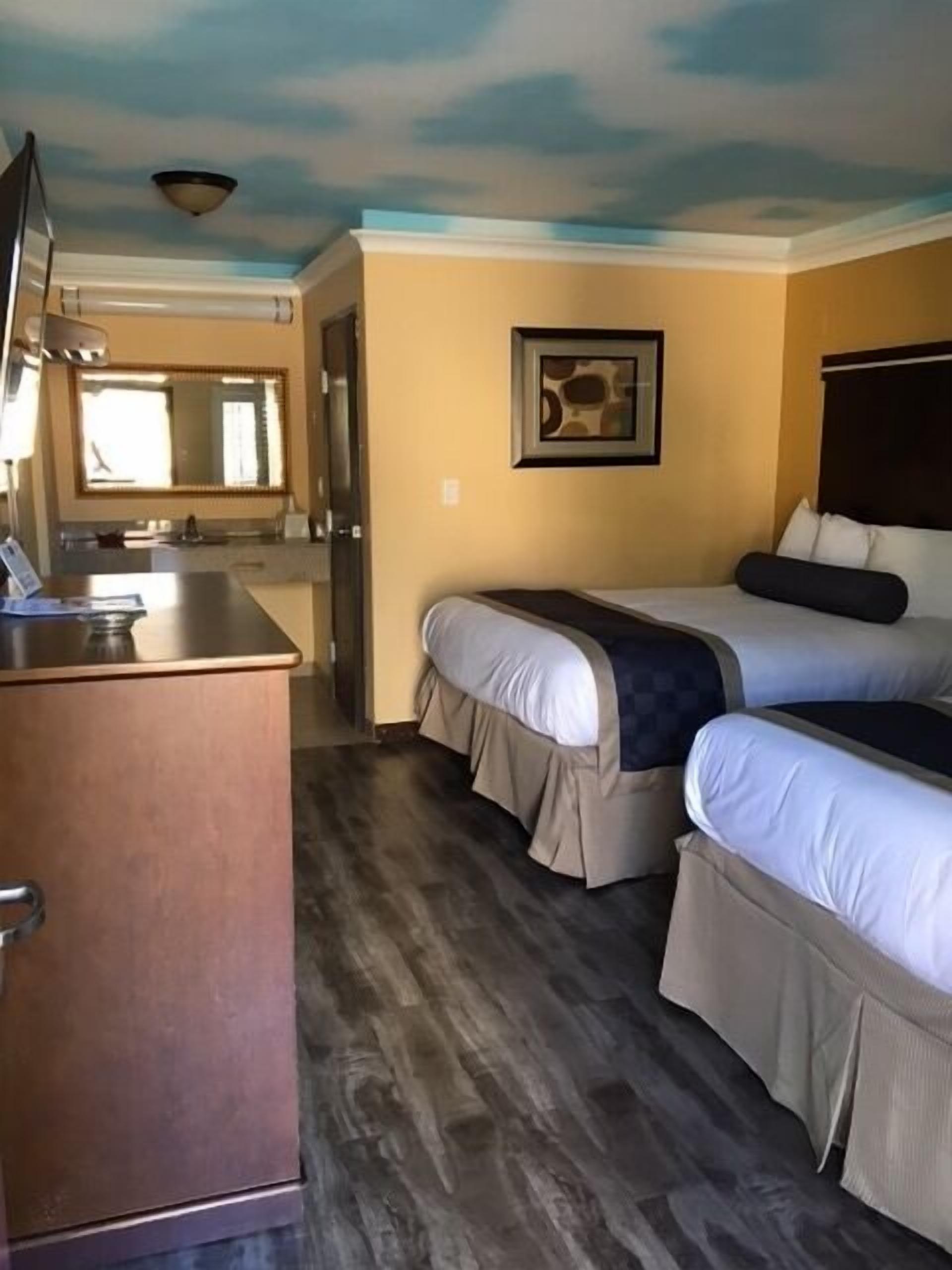 A room with two beds, a kitchenette, a mirror, and a ceiling with a cloud design.