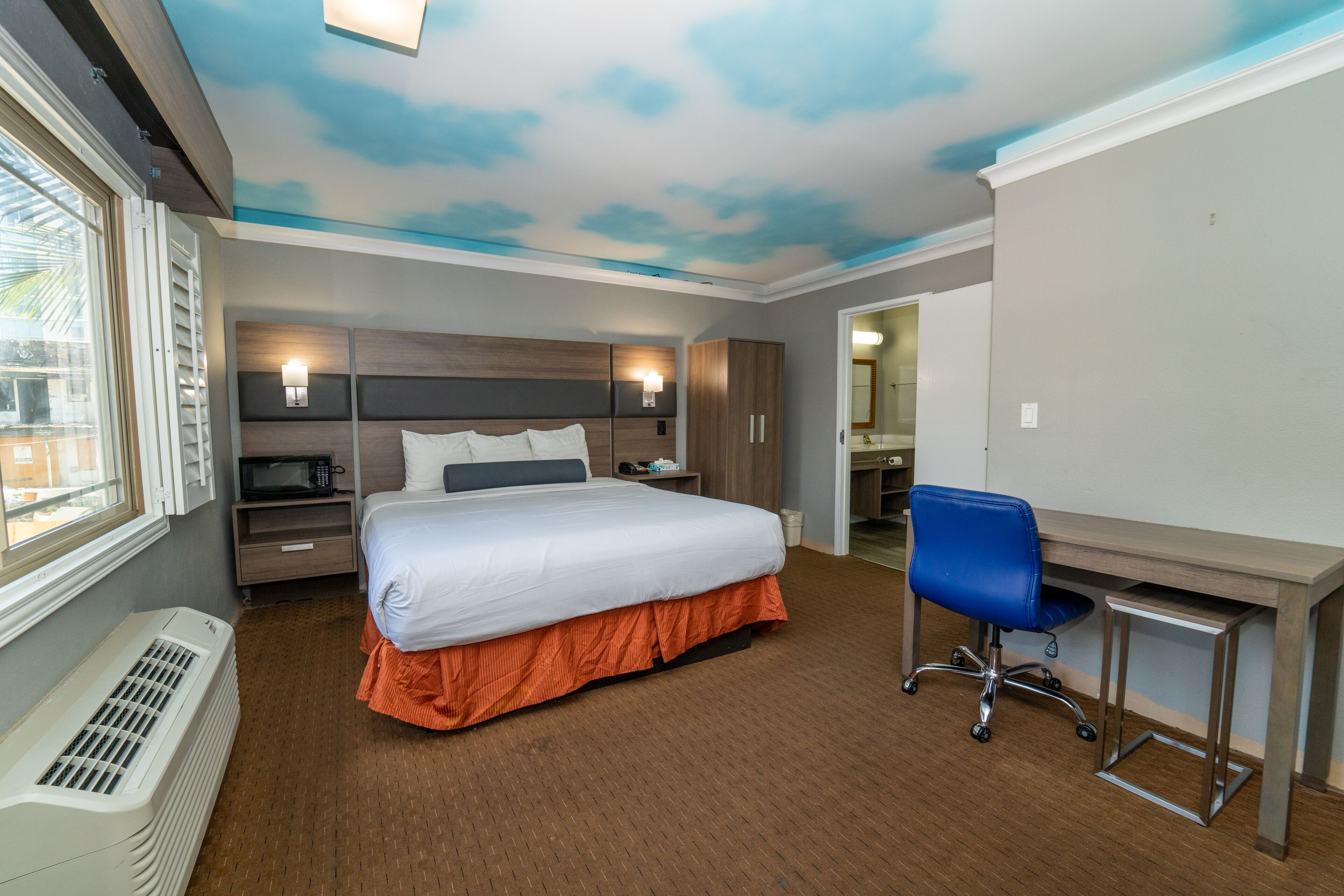 A hotel room with a bed, a desk, a chair, a microwave, and a heater.