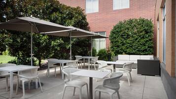 Un patio al aire libre con mobiliario de mimbre blanco, incluyendo mesas y sillas, y un gran paraguas que da sombra.
