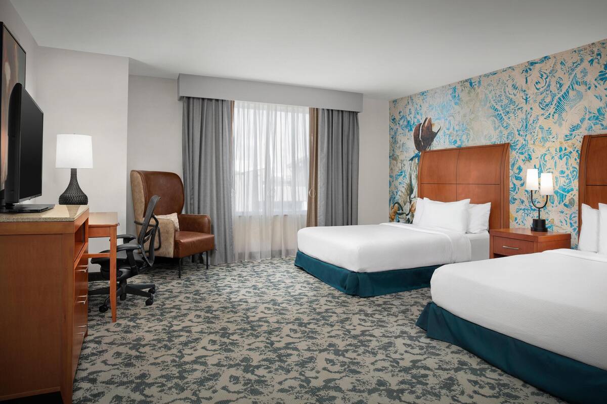 A hotel room with two beds, a desk, a chair, a TV, and a window with curtains.