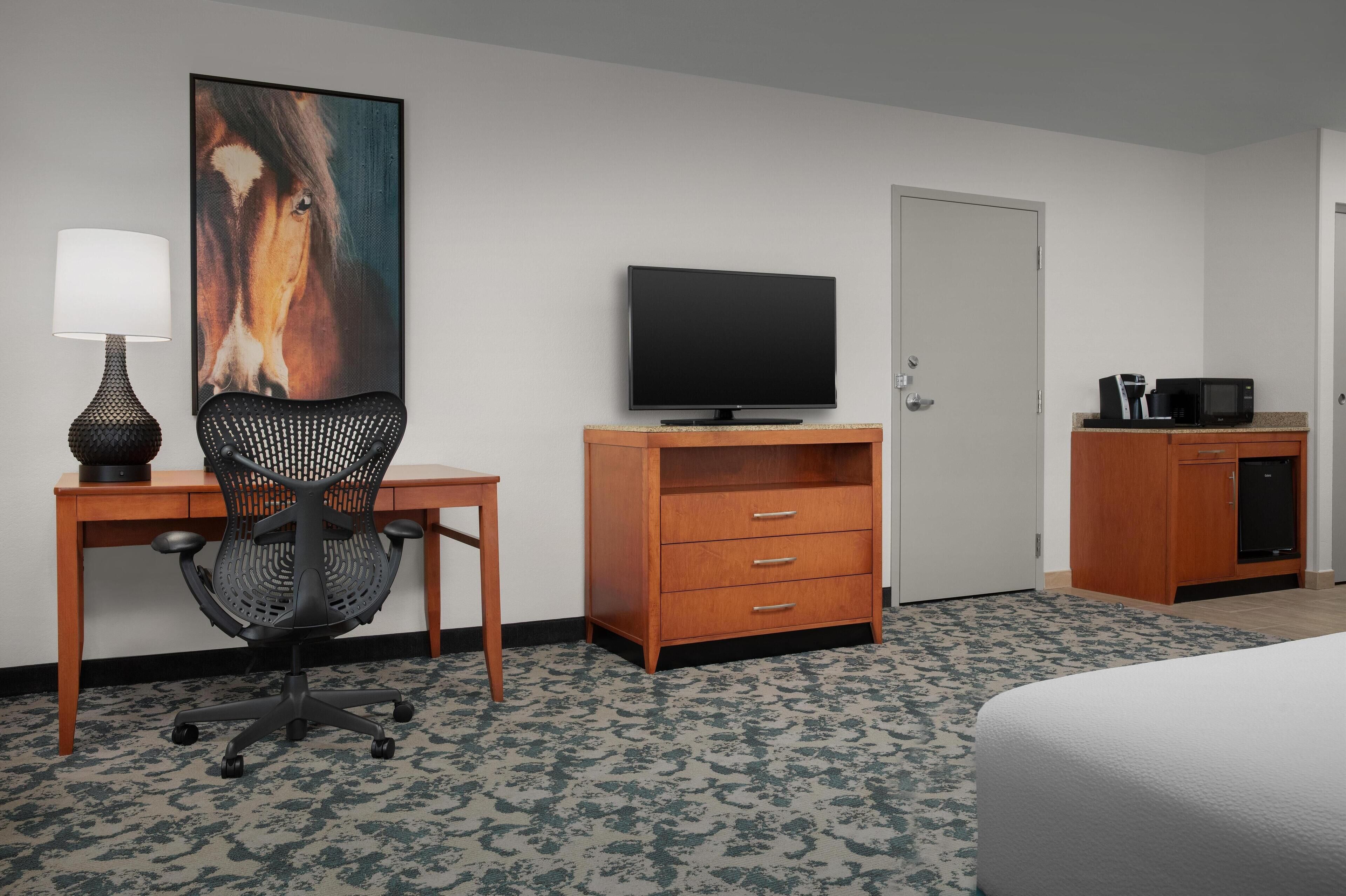 A hotel room with a bed, a desk with a lamp, a television, a wooden dresser, and a microwave.
