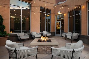 An outdoor seating area with wicker chairs, a central fire feature, and string lights. - Hilton Garden Inn Fort Worth Medical Center (Fort Worth)