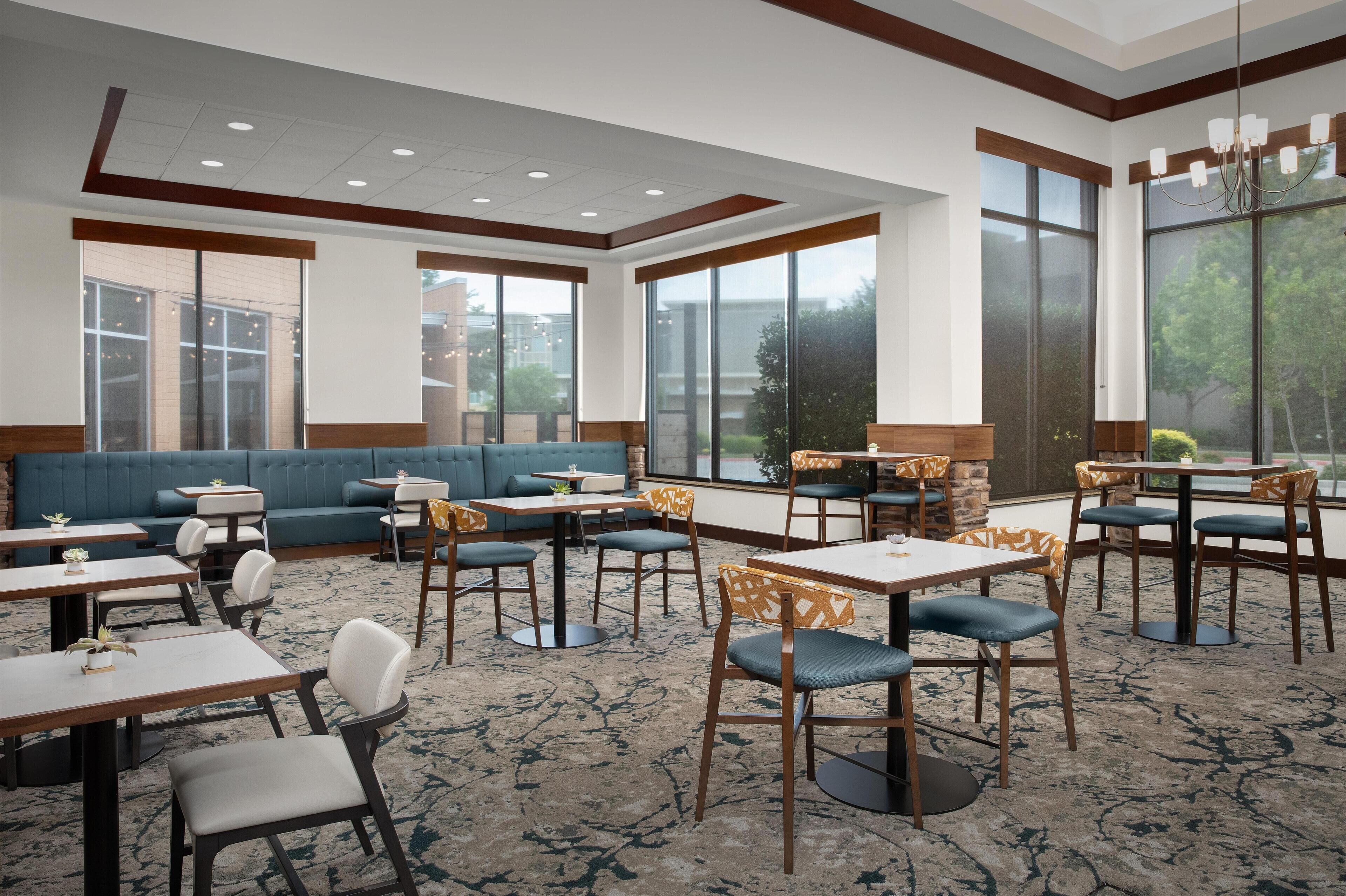 A hotel dining area with tables, chairs, a sofa, and large windows.
