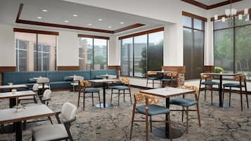 A hotel dining area with tables, chairs, a sofa, and large windows.