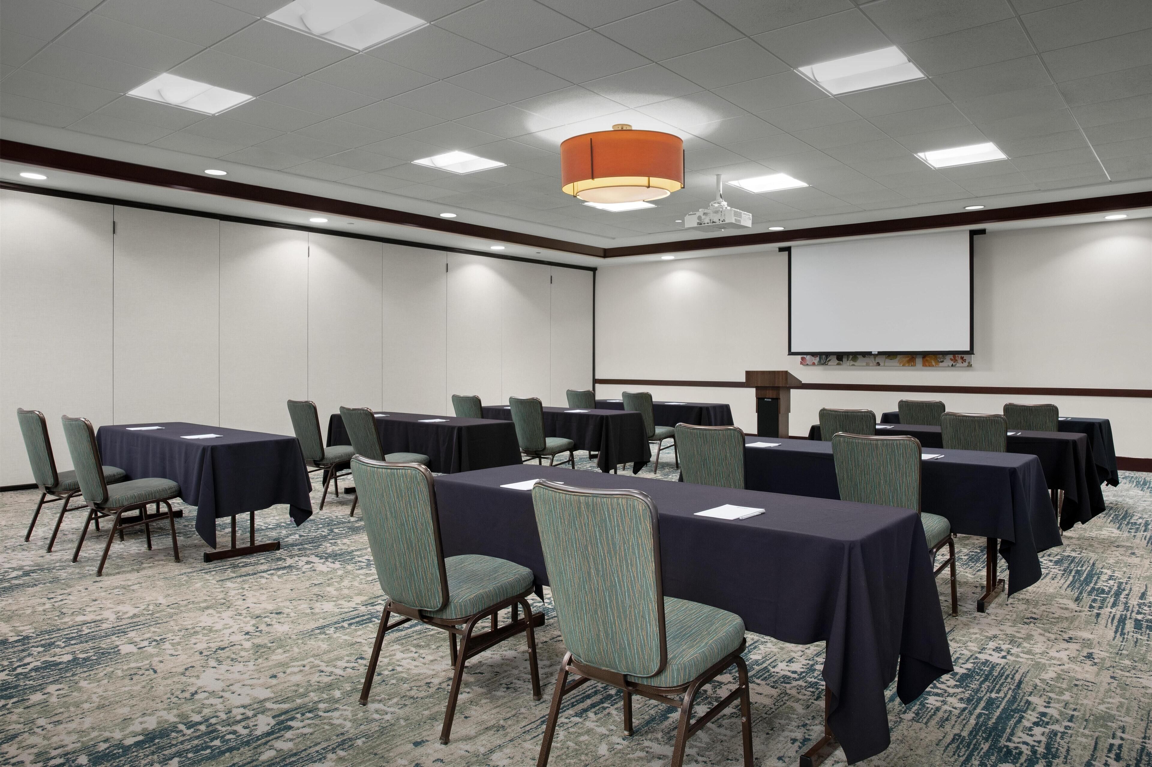 A conference room with tables and chairs, a projector screen, and a carpeted floor.