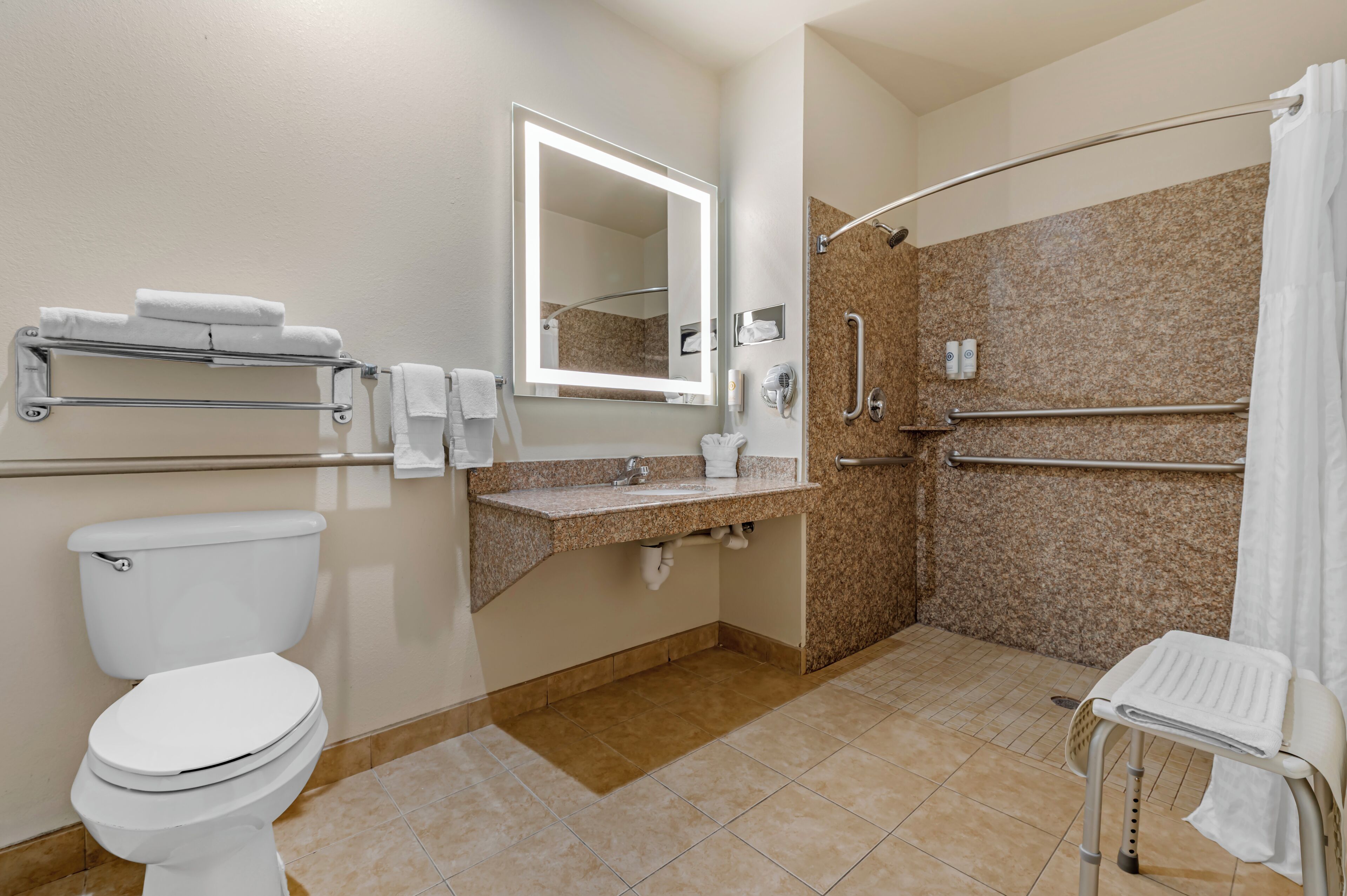Une salle de bain équipée de toilettes, d’un lavabo, d’un miroir, d’un porte-serviettes, d’une douche avec des barres d’appui et d’un banc accessible aux personnes en fauteuil roulant.