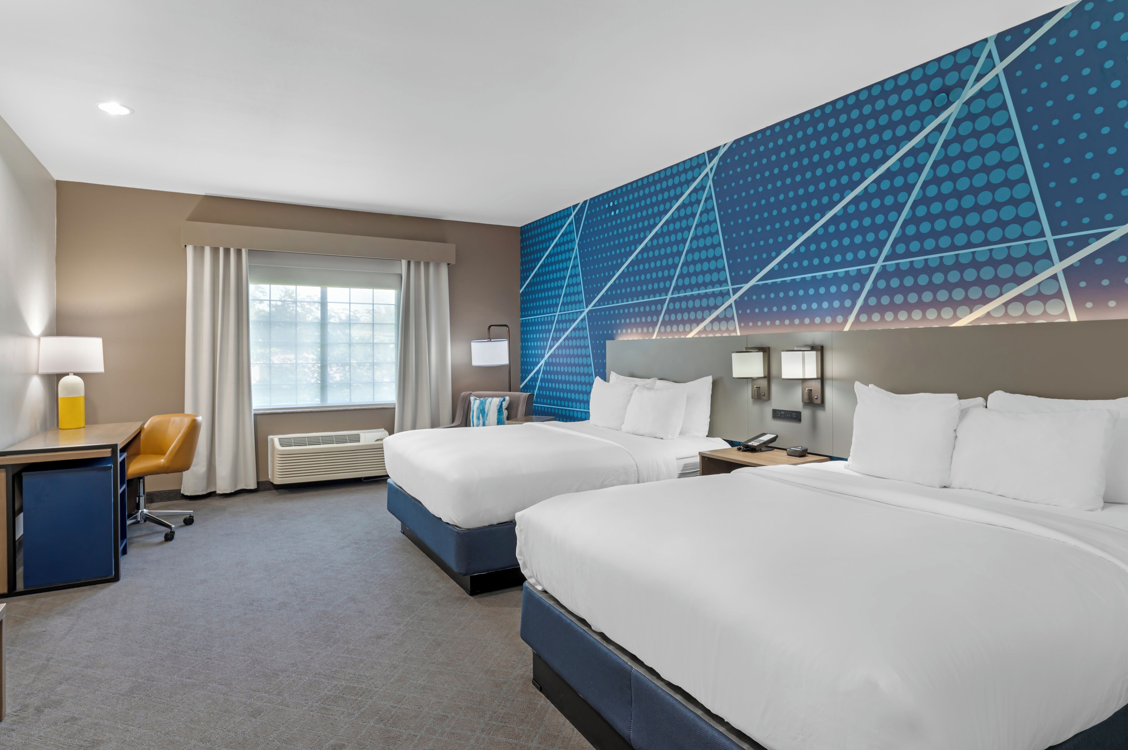 A hotel room with two beds, a desk, a chair, a window with curtains, and a blue patterned wall.