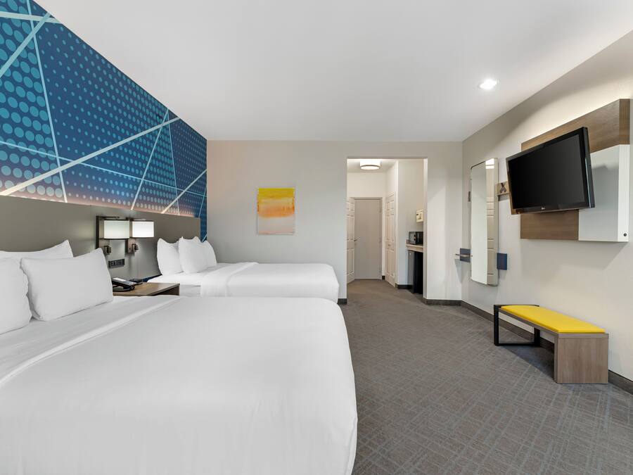 A hotel room with a large bed, a TV mounted on the wall, a bench, and a hallway leading to a bathroom.