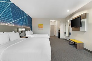 Habitación de hotel con una cama grande, televisión montada en la pared, un banco y un pasillo que lleva a un baño.