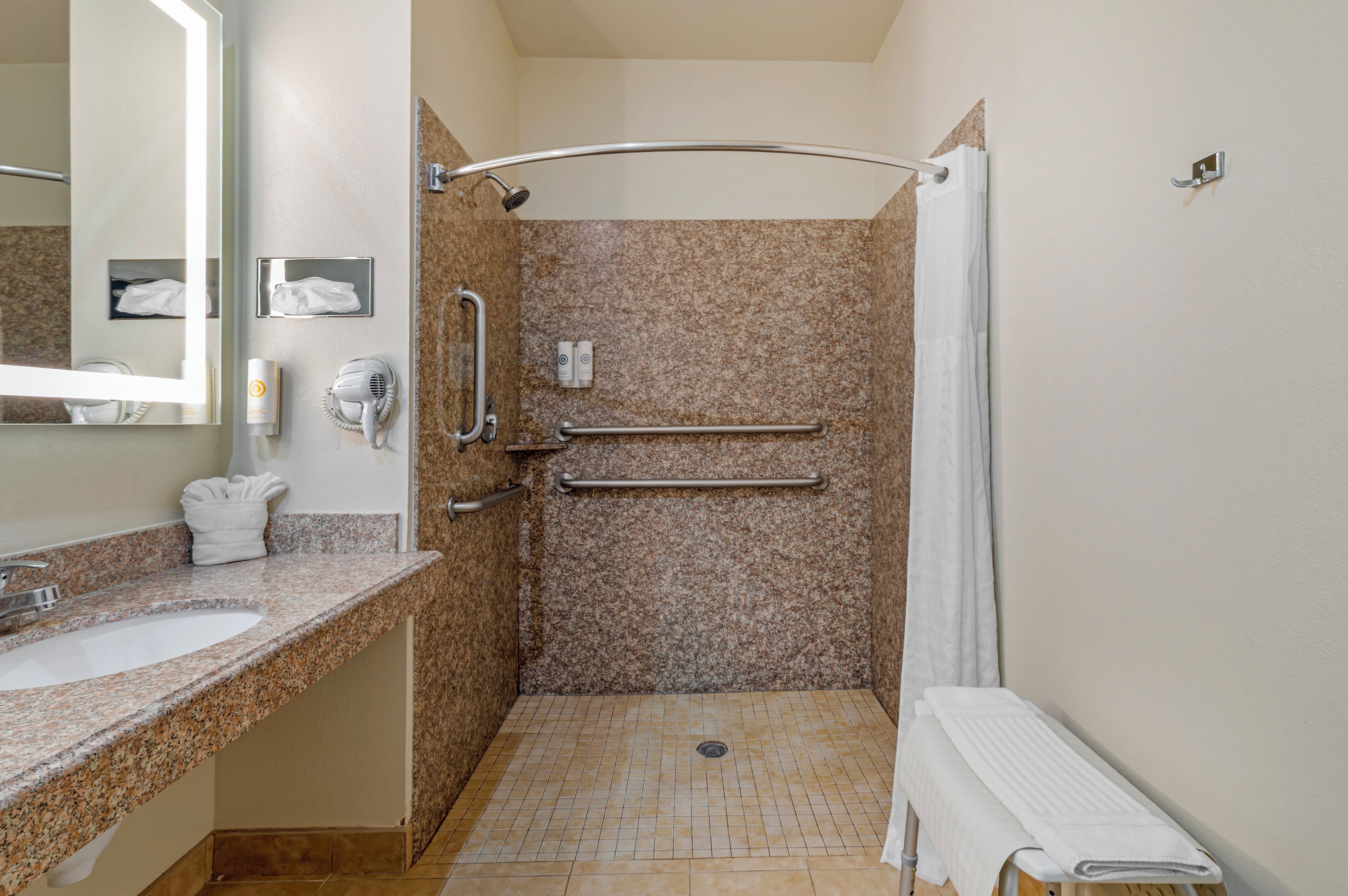 A bathroom with a sink, mirror, towel rack, hand dryer, and a shower with a grab bar.